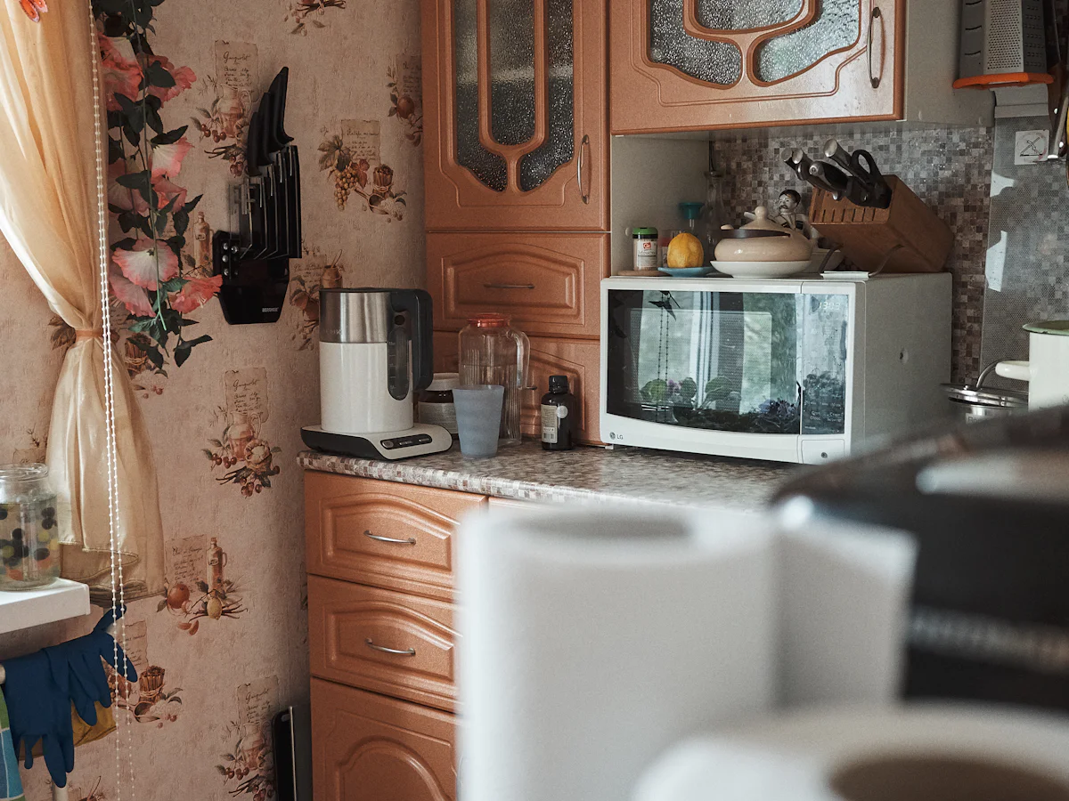 White microwave oven on a kitchen counter under wooden wall cabinets, with a kettle and paper towel rolls nearby
